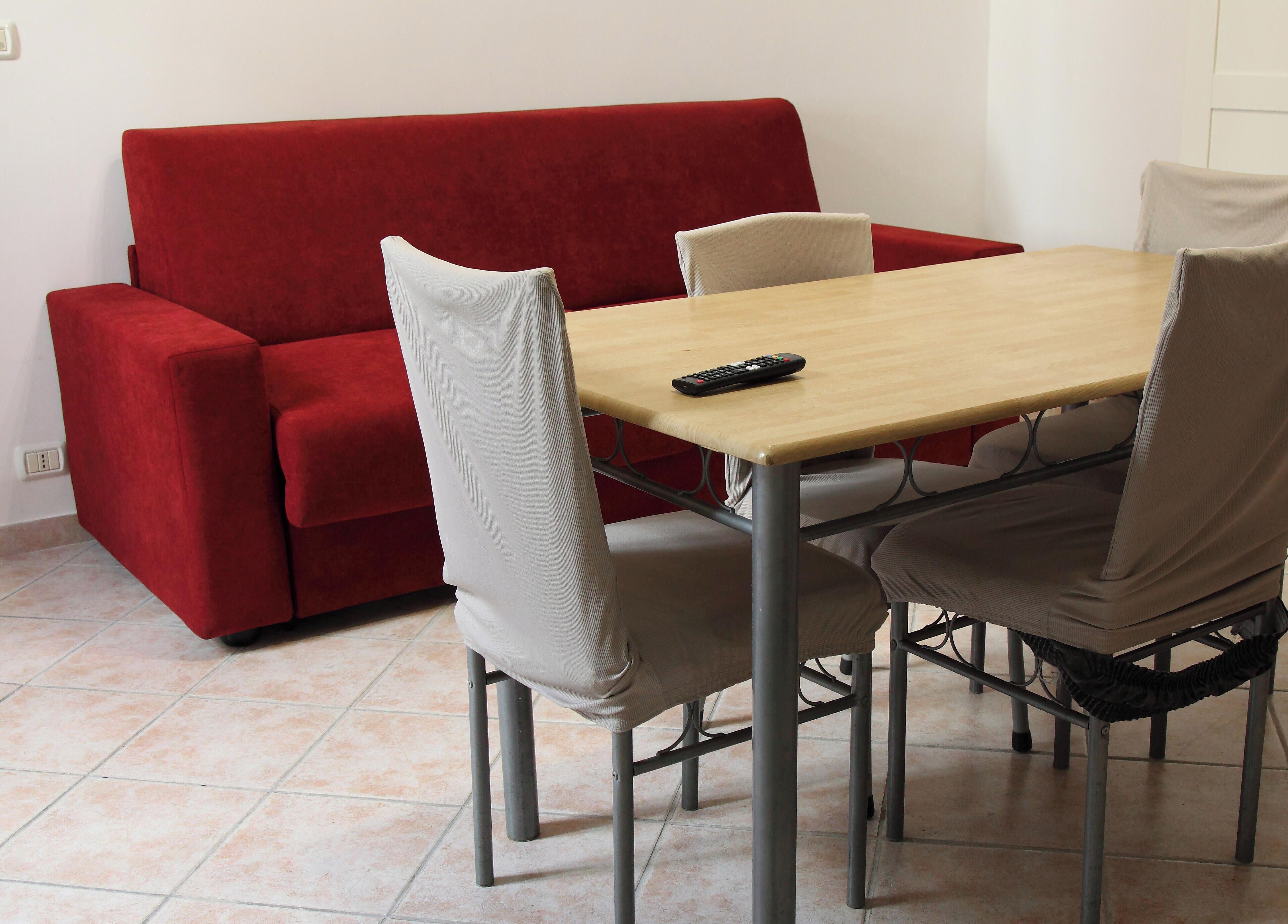 Un comedor con un sofá rojo, una mesa de madera y cuatro sillas. Un control remoto está sobre la mesa.