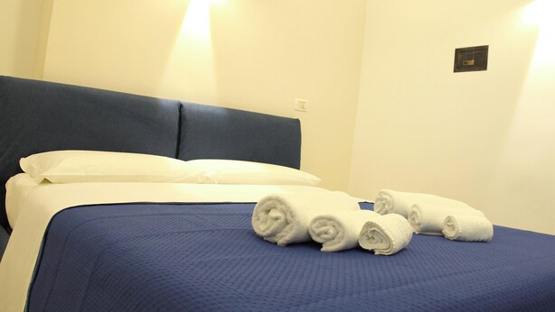 Una cama bien hecha con una colcha azul y almohadas blancas, junto con toallas blancas enrolladas sobre la cama.