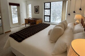 Ein Schlafzimmer mit einem großen Bett, einem Schreibtisch und einem Stuhl. Fenster bieten Blick auf Bäume.