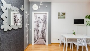Een moderne kamer met een houten vloer, een witte eettafel met stoelen, een flatscreen televisie en een decoratieve spiegel aan de muur.
