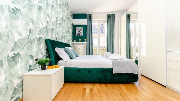 Een moderne slaapkamer met een met groen bekleden bed, een witte nachtkastje en een muur met een bladmotief.
