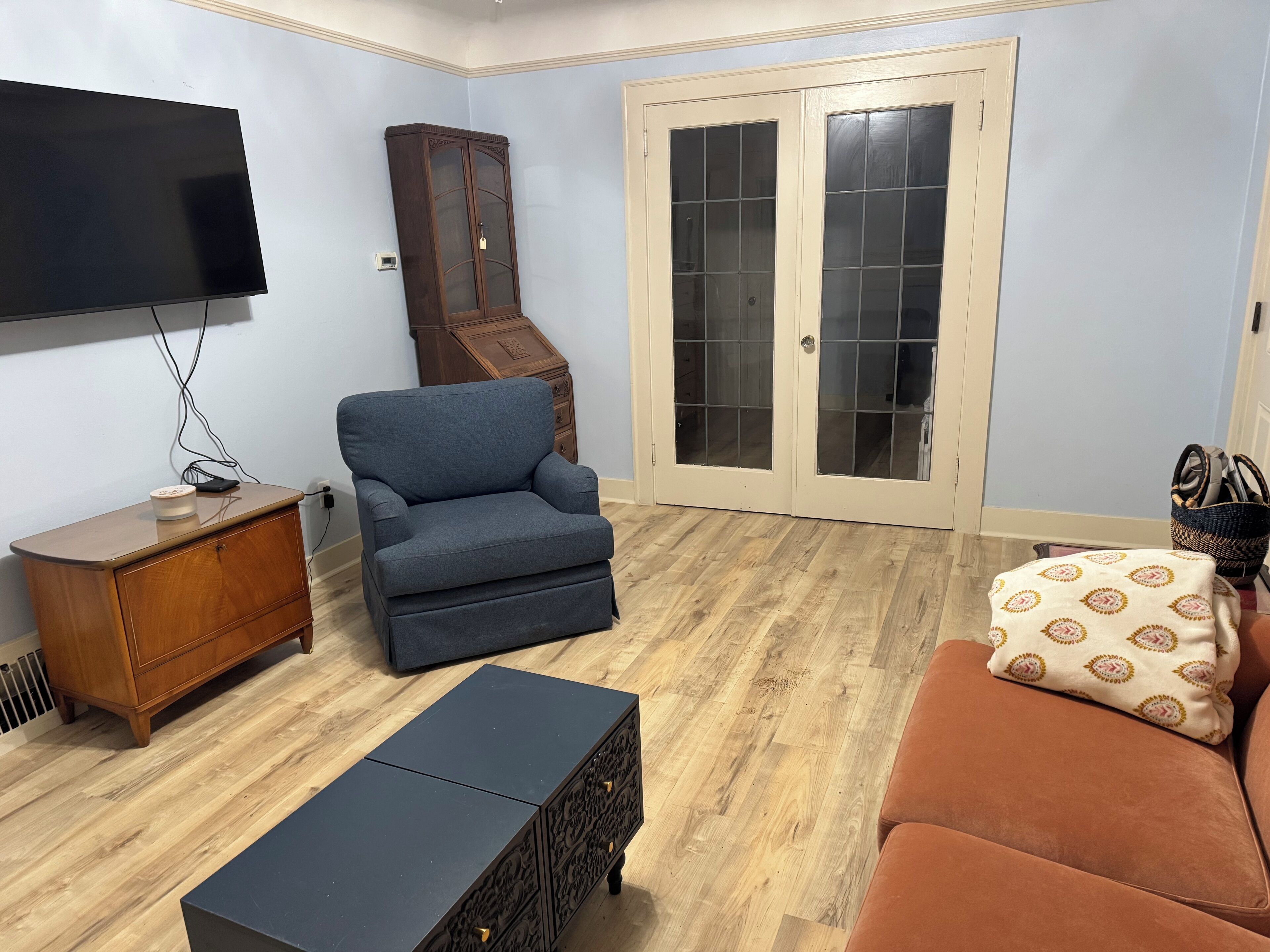 A living room with a flat-screen TV, a blue armchair, a wooden cabinet, a black coffee table, a brown sofa, and a patterned throw pillow.