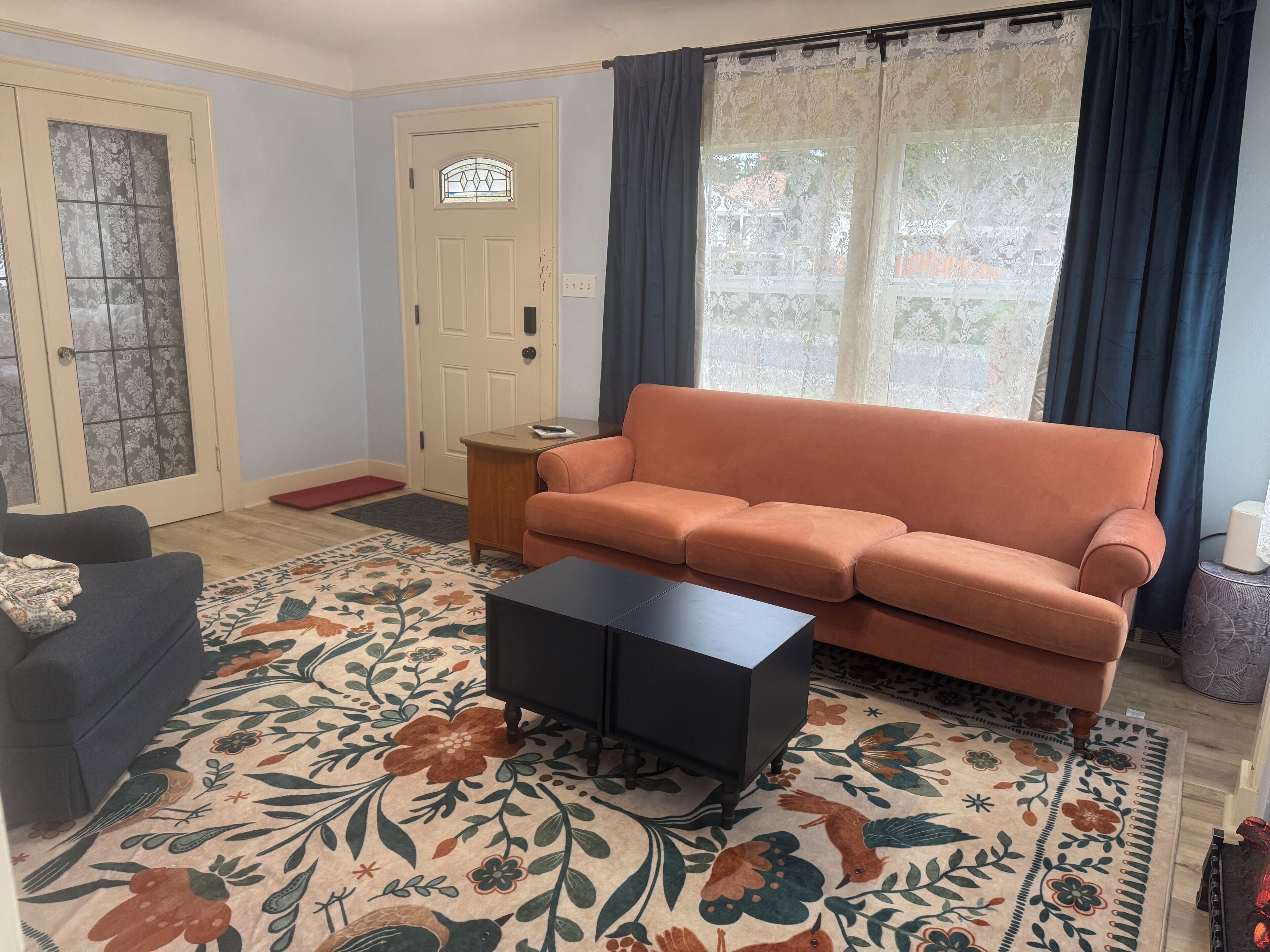 A living room with a sofa, a coffee table, a patterned rug, a door with a stained glass window, and a window with curtains.