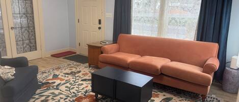 A living room with a sofa, a coffee table, a patterned rug, a door with a stained glass window, and a window with curtains.
