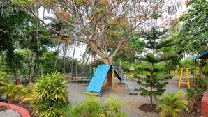 Un parc infantil amb tobogan blau i cadira d'osc lleure groga, envoltat de plantes en testos i arbres.