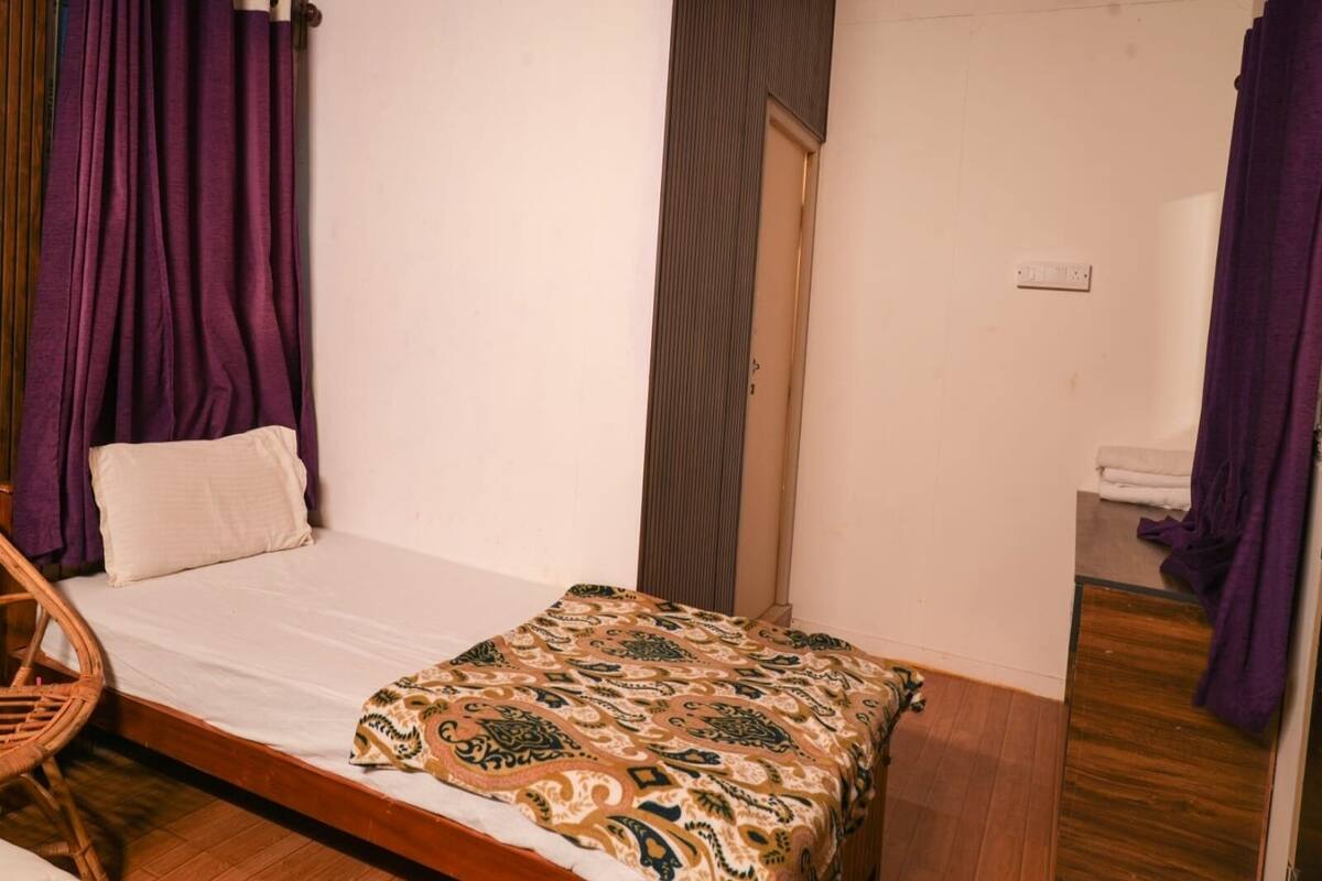 Una habitación pequeña con una cama individual, suelo de madera, colcha estampada y cortinas moradas.