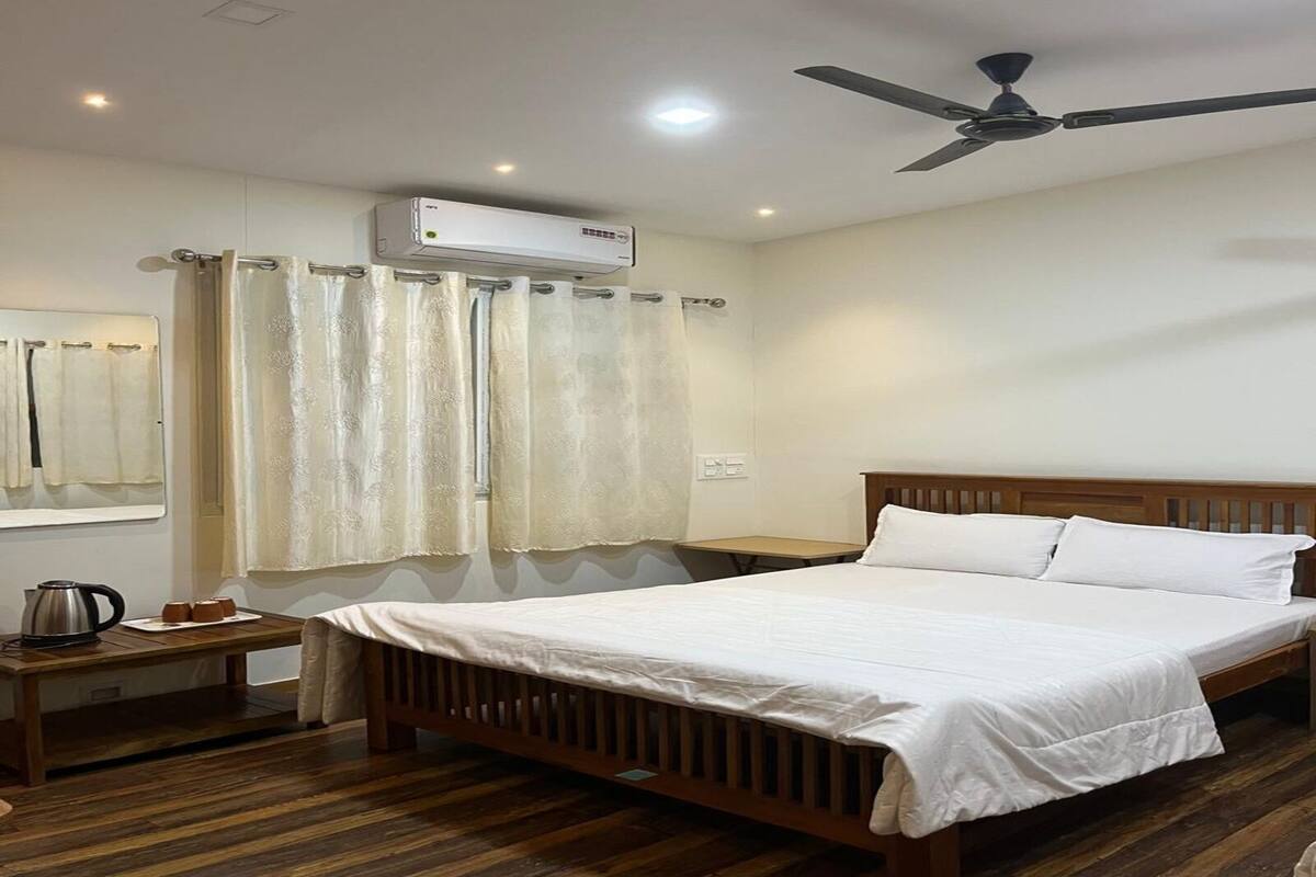 Un dormitorio con cama de madera, ropa de cama blanca, ventilador de techo, equipo de aire acondicionado, una mesita con tetera y un espejo en la pared.