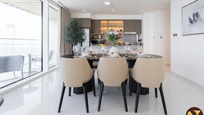 A modern dining area with a large window, a potted plant, and a kitchen in the background. - Vogue| Grand Blue Beachfront Designed By Elie Saab (Dubai)