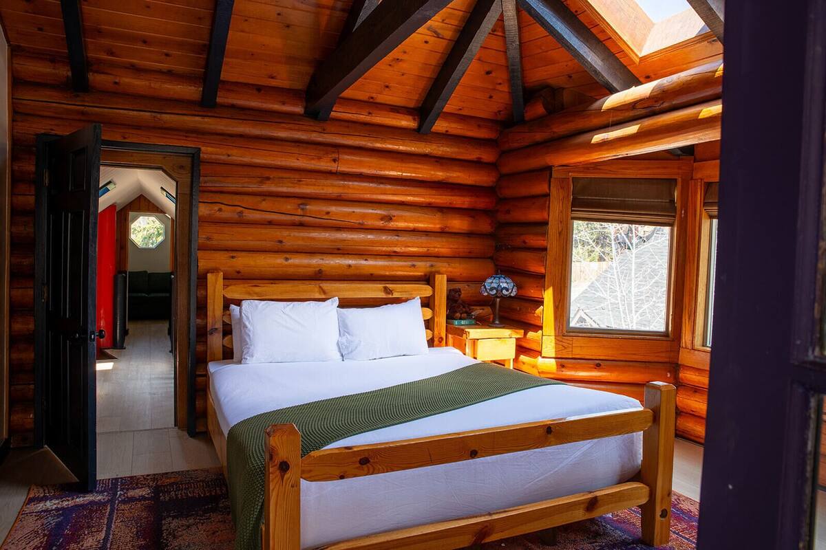 A log cabin bedroom with a wooden bed, a nightstand, a window, and a door leading to another room.