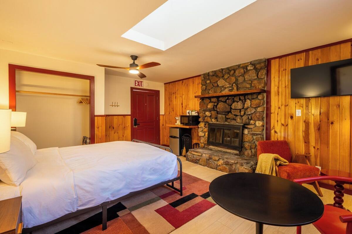 A hotel room with a bed, a small table, a chair, a stone fireplace, a television, and a ceiling fan.
