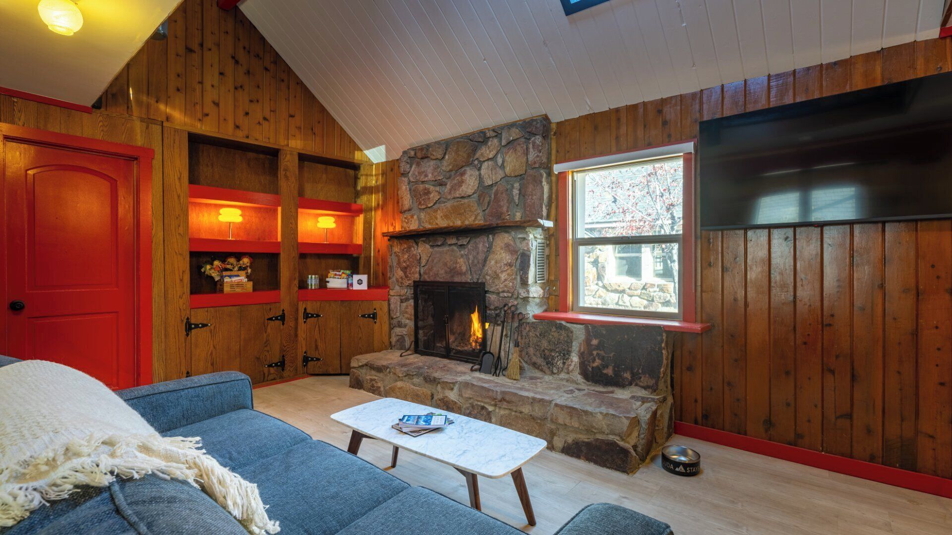 A cozy living room with a stone fireplace, wooden walls, a red door, and a window with a view.