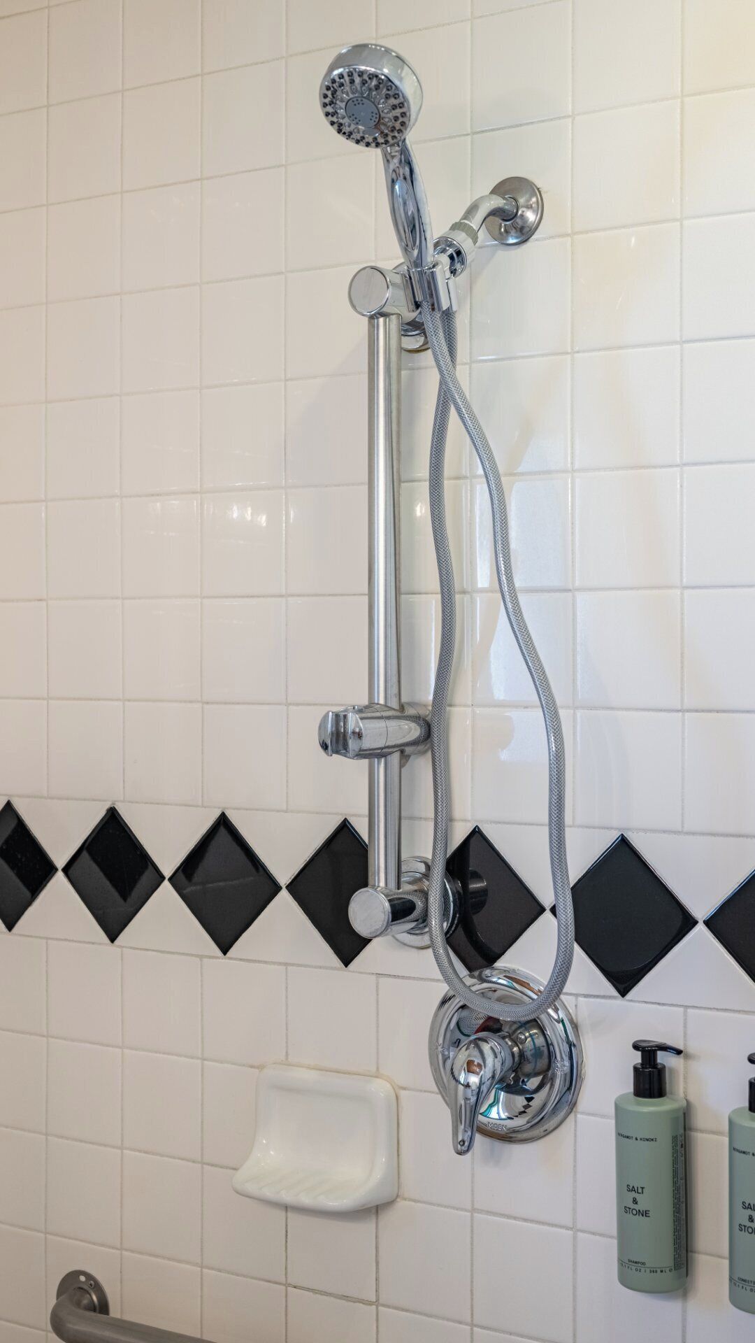 A shower with a handheld showerhead and a flexible hose. There is a soap dish and two bottles of shampoo on the wall.