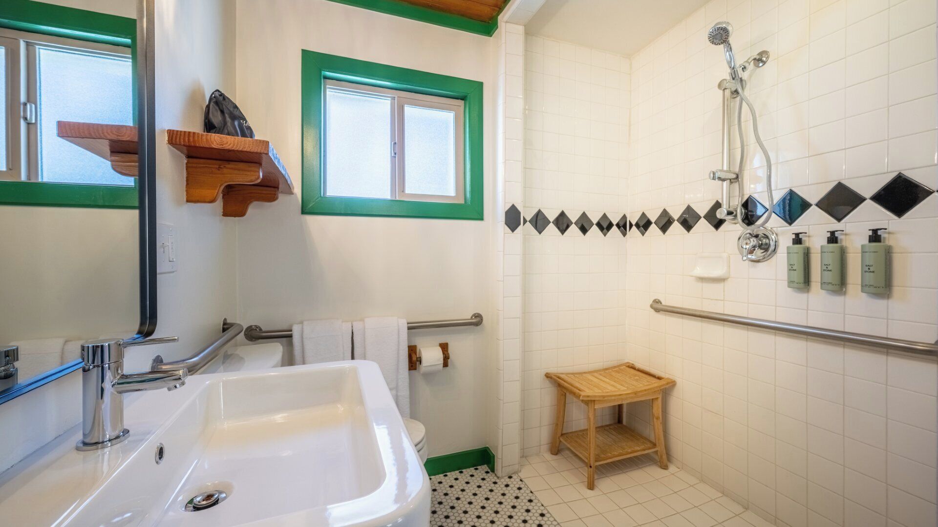 Une salle de bain avec un lavabo blanc, une douche équipée d’un pommeau de douche à main, un petit tabouret en bois et une fenêtre avec des bordures vertes.