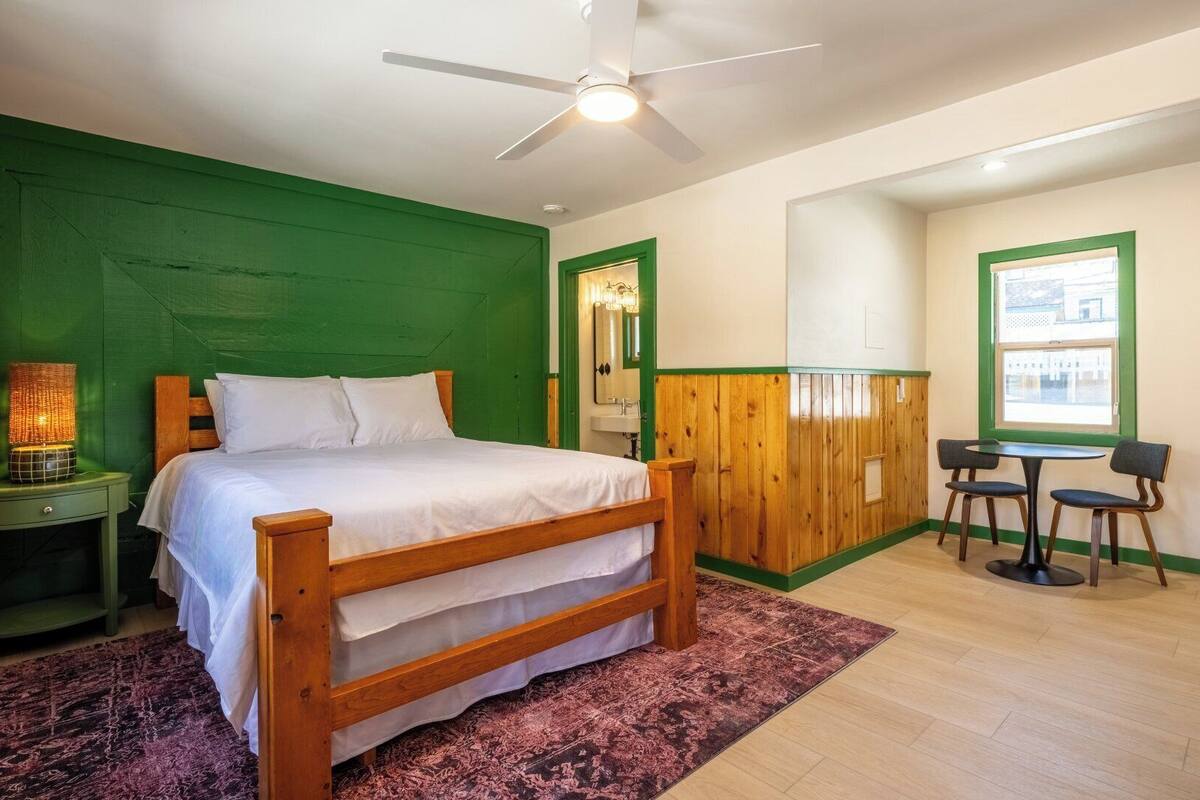 A bedroom with a bed, a nightstand, a green wall, a wooden partition, a window, and a table with chairs.