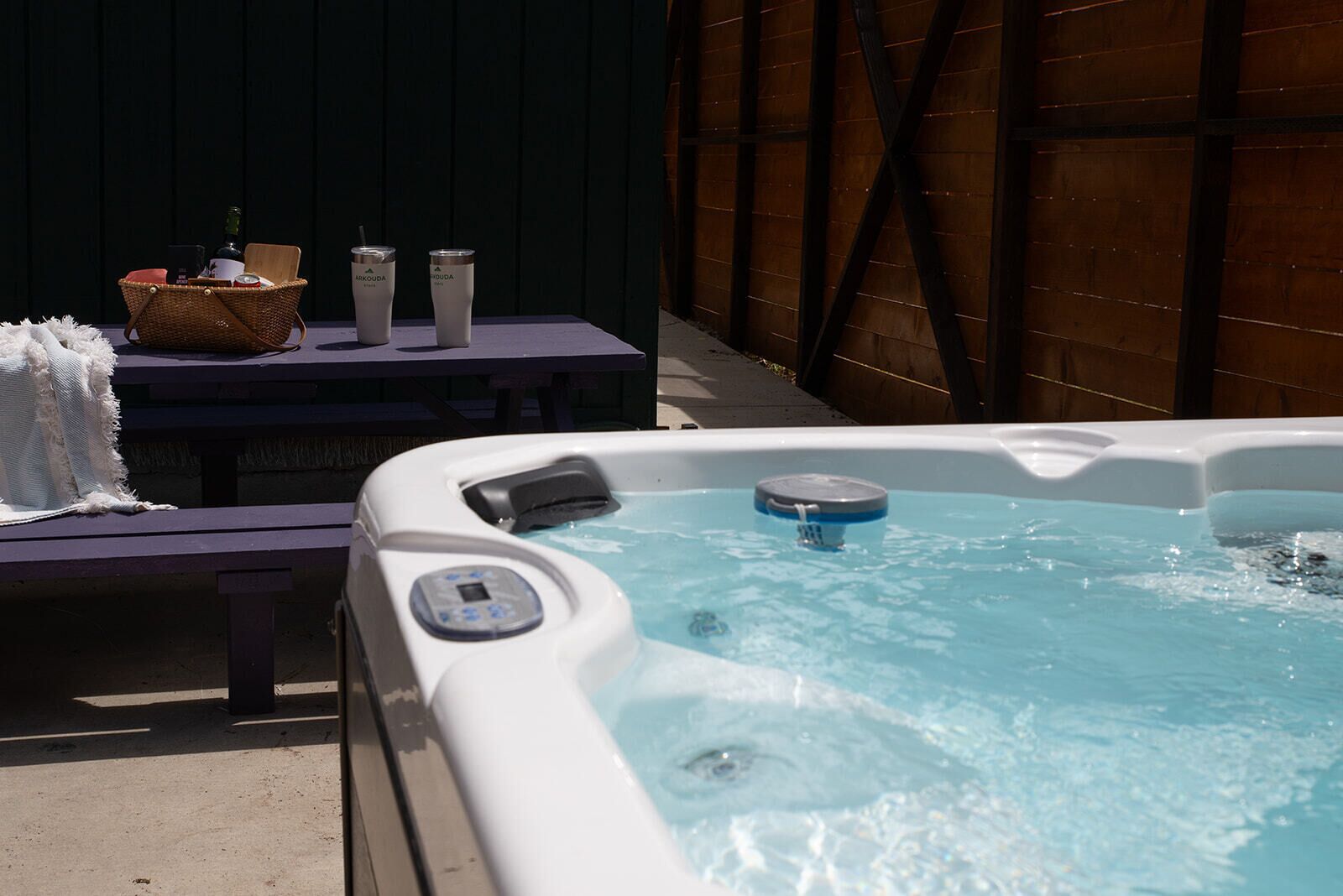 A hot tub with a control panel, a wicker basket, and a table with cups and a bottle.