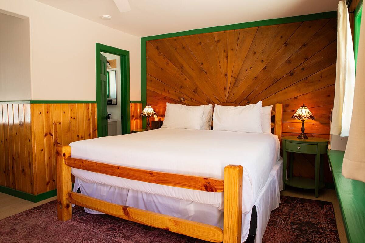 A wooden bed with white bedding, a nightstand with a lamp, and a green-framed mirror.