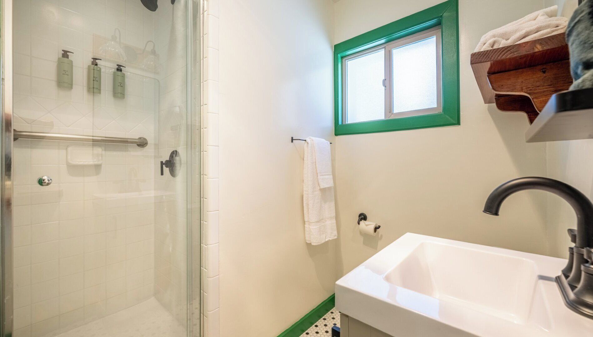A bathroom with a glass shower enclosure, a white sink, a window with a green frame, and a wooden shelf with towels.