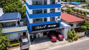 Un edificio de varios pisos con fachada azul y blanca, identificado como "Collection O JM Lodge", que cuenta con un estacionamiento y varios automóviles.