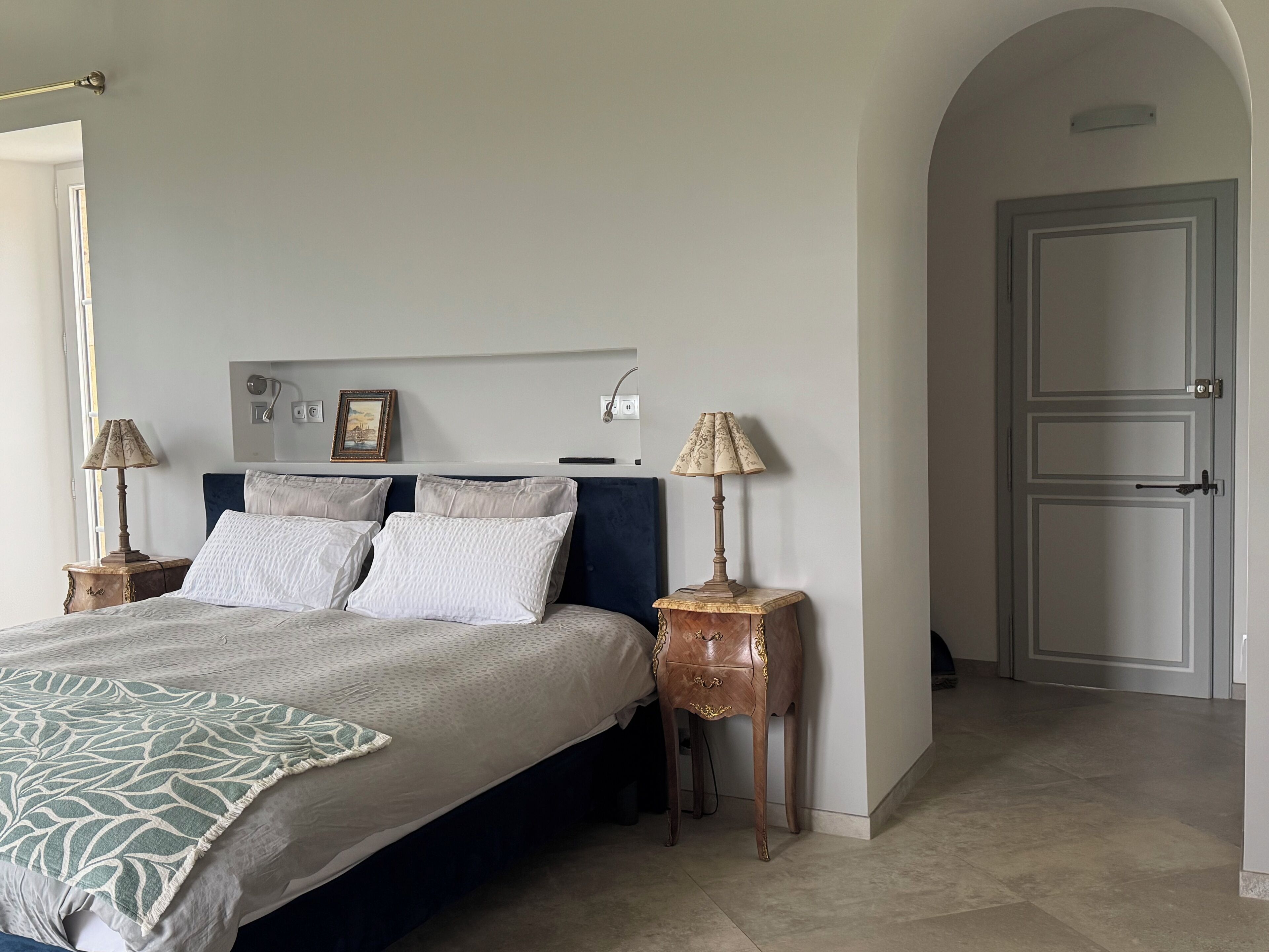 Un dormitorio con una cama, dos mesitas de noche, una lámpara de pared, un cuadro enmarcado y una puerta.