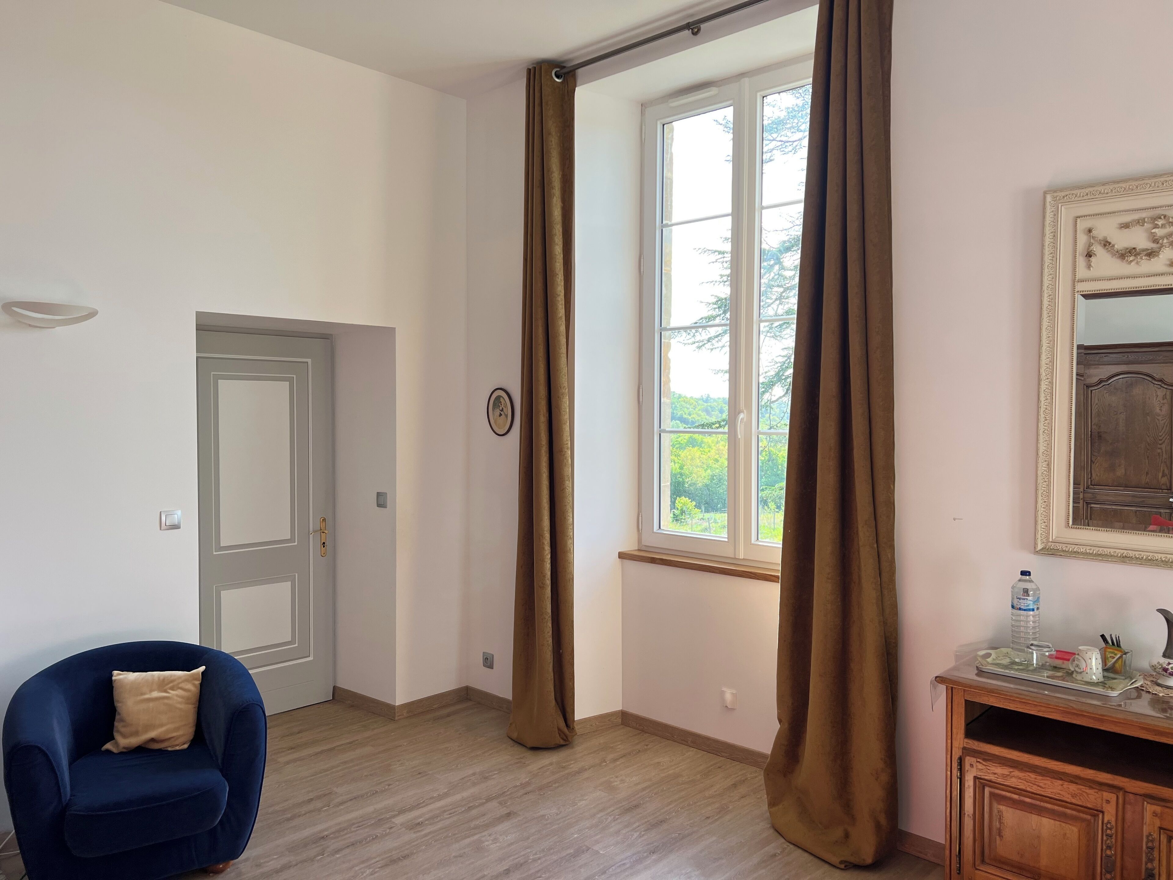 A room with a door, a window with curtains, a chair, a wooden cabinet, and a wall-mounted light fixture.