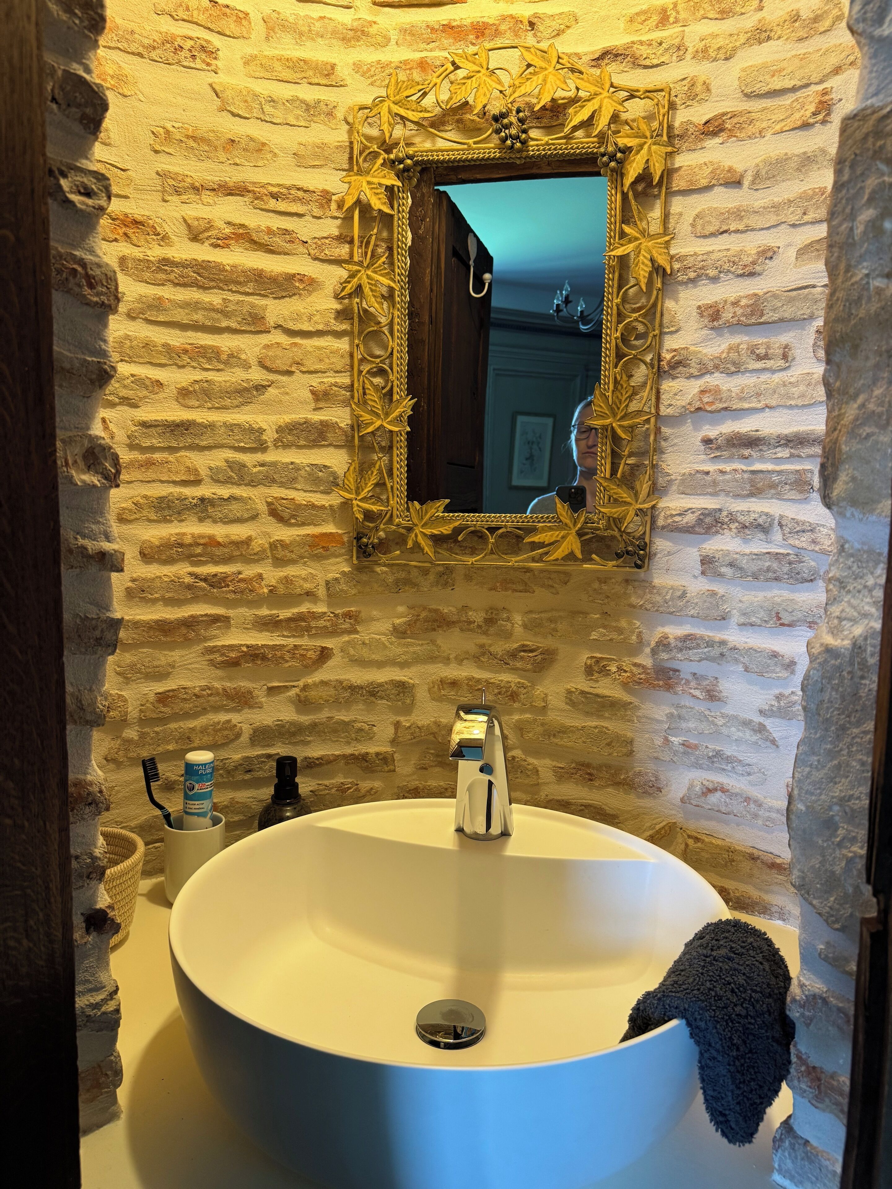 Un baño con pared de piedra, un lavabo redondo, un espejo con marco dorado y una toalla colgada en la pared.