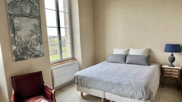 A bedroom with a bed, a chair, a nightstand, a lamp, a window, and a wall-mounted picture.