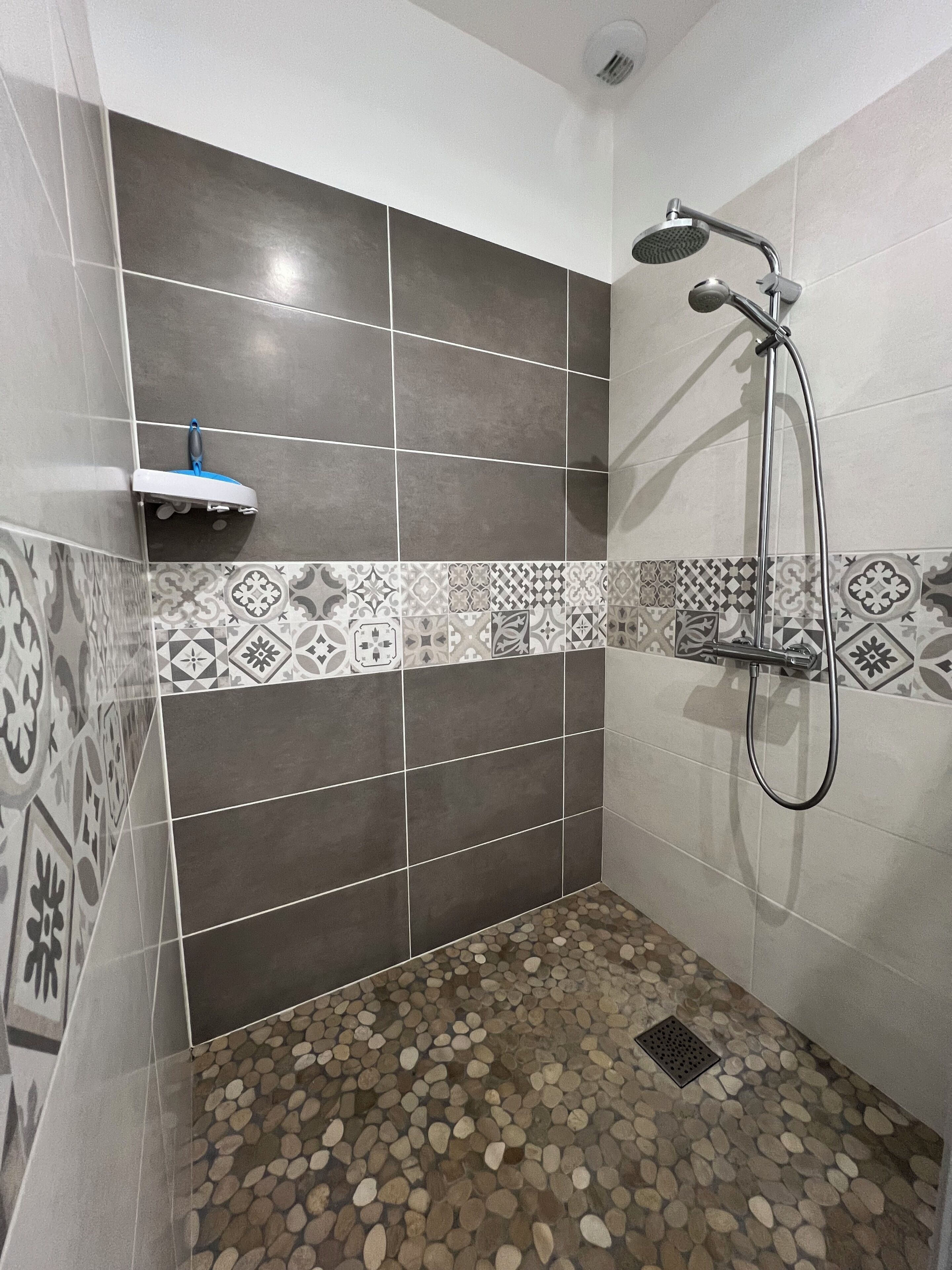 A shower with a handheld showerhead, a tiled floor with pebble patterns, and a shelf with a blue object.