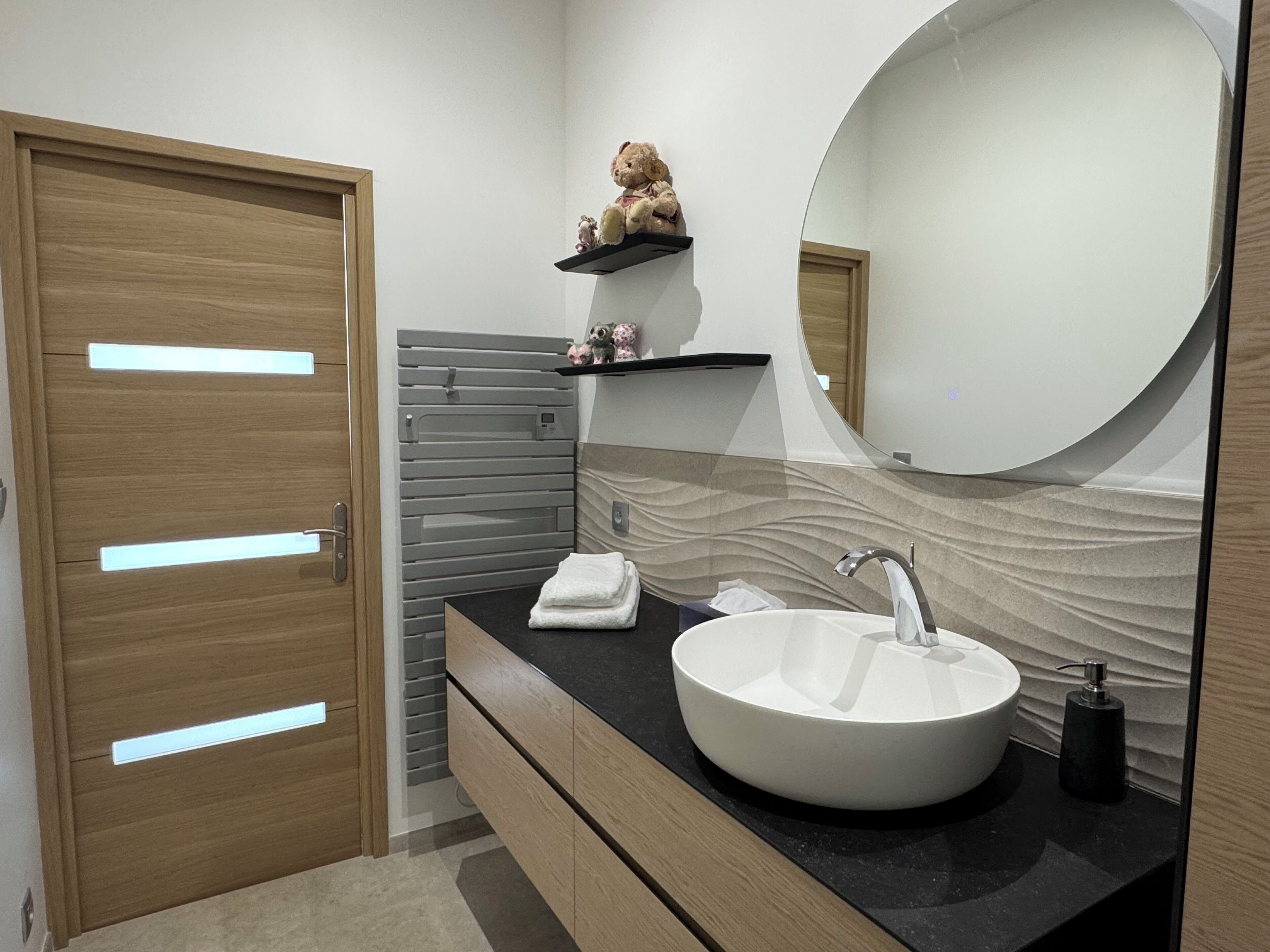 Un baño moderno con puerta de madera, un lavamanos blanco, un espejo y un perchero con detalles decorativos.