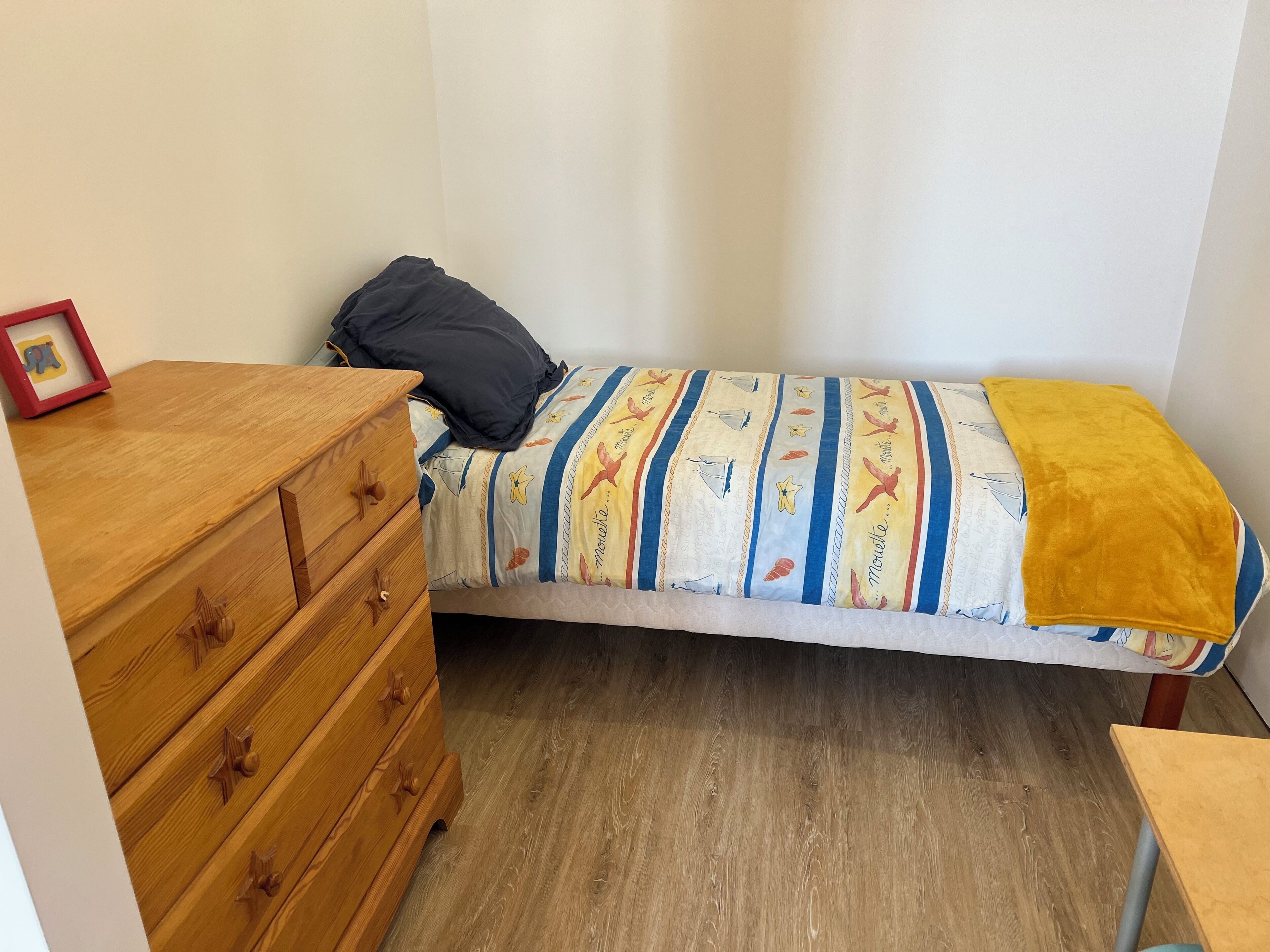 Une petite chambre avec un lit simple, une commode en bois, une table de chevet et une couvre-lit aux couleurs vives.