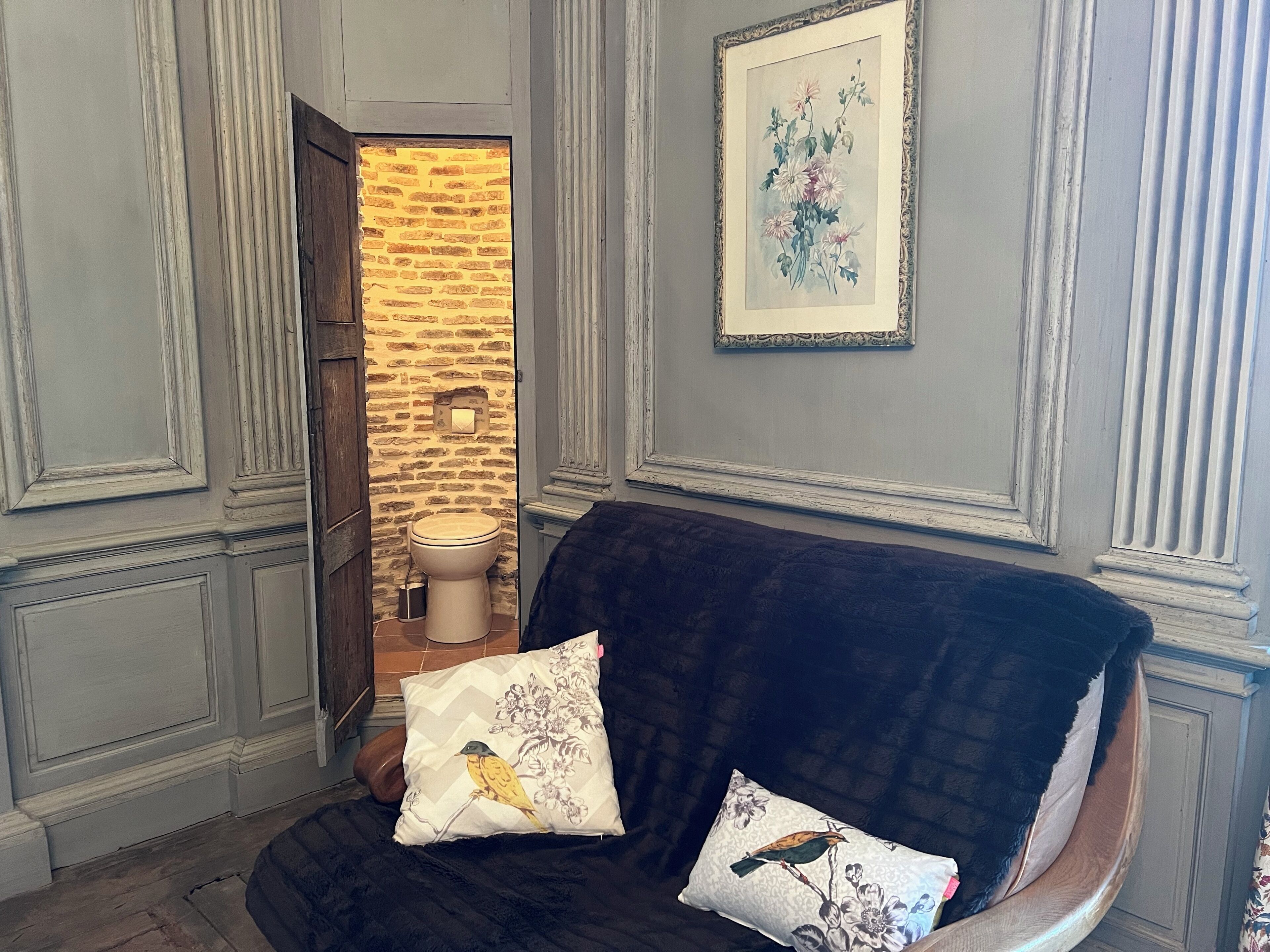 Ein Zimmer mit einem dunklen Sofa, zwei Zierkissen, einem gerahmten Blumenbild und Blick in ein Badezimmer mit Ziegelwand und Toilette.