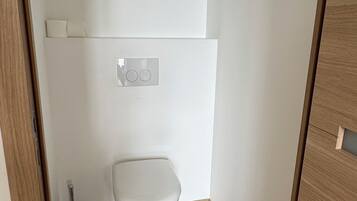 A small, modern bathroom with a white toilet, a flush handle, and a plunger on the floor.