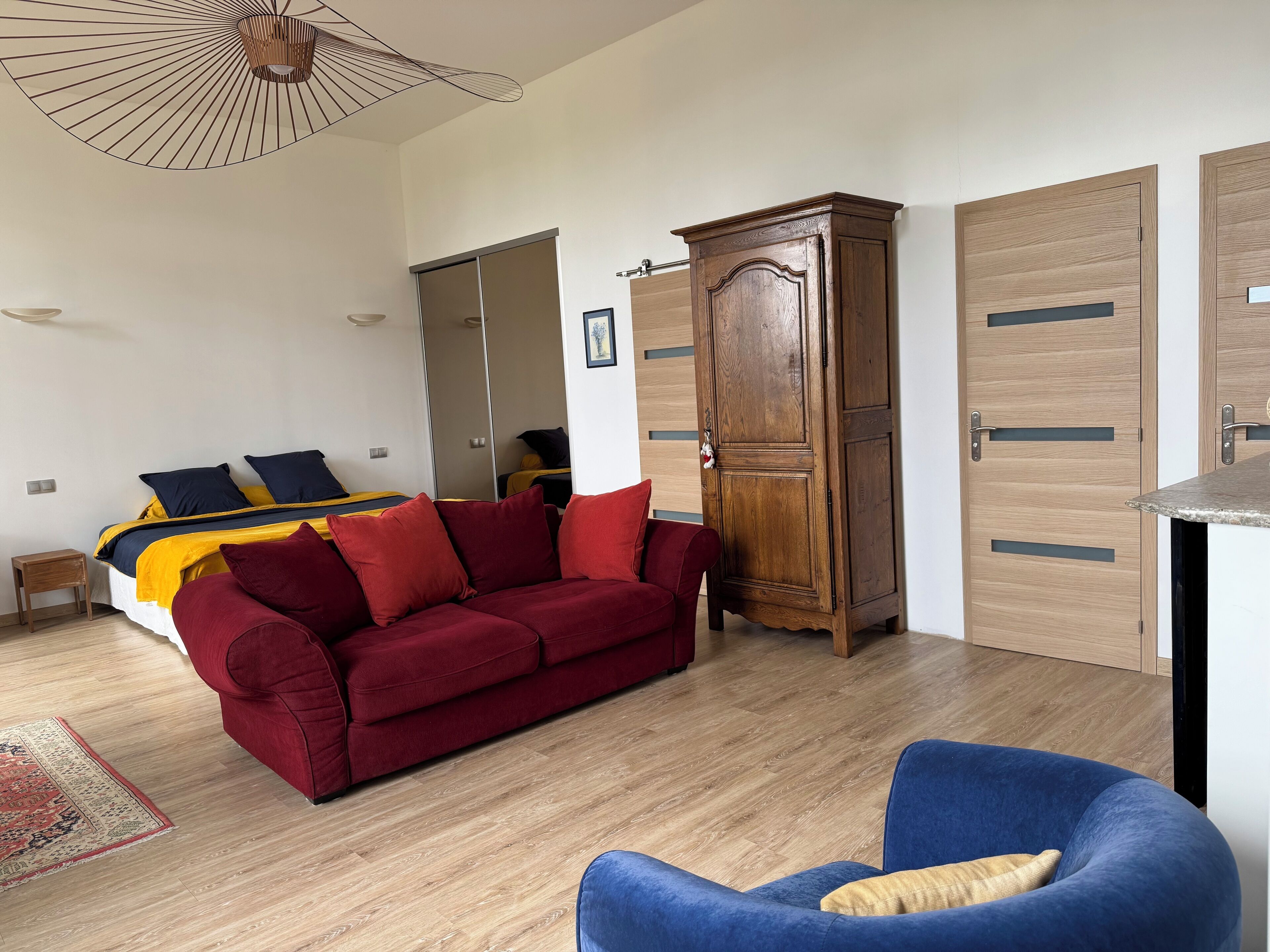 Una habitación con un sofá rojo, un sillón azul, un armario de madera y una cama con ropa de cama amarilla y azul.