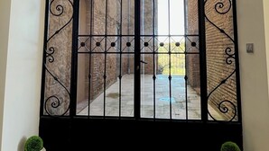 Una reja de hierro forjado negro con un diseño decorativo, que da acceso a un patio con un muro de ladrillo y un cielo azul despejado.