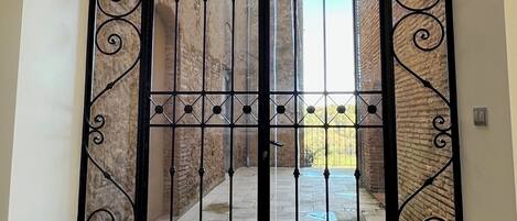 A black wrought iron gate with a decorative design, leading to a courtyard with a brick wall and a clear blue sky.