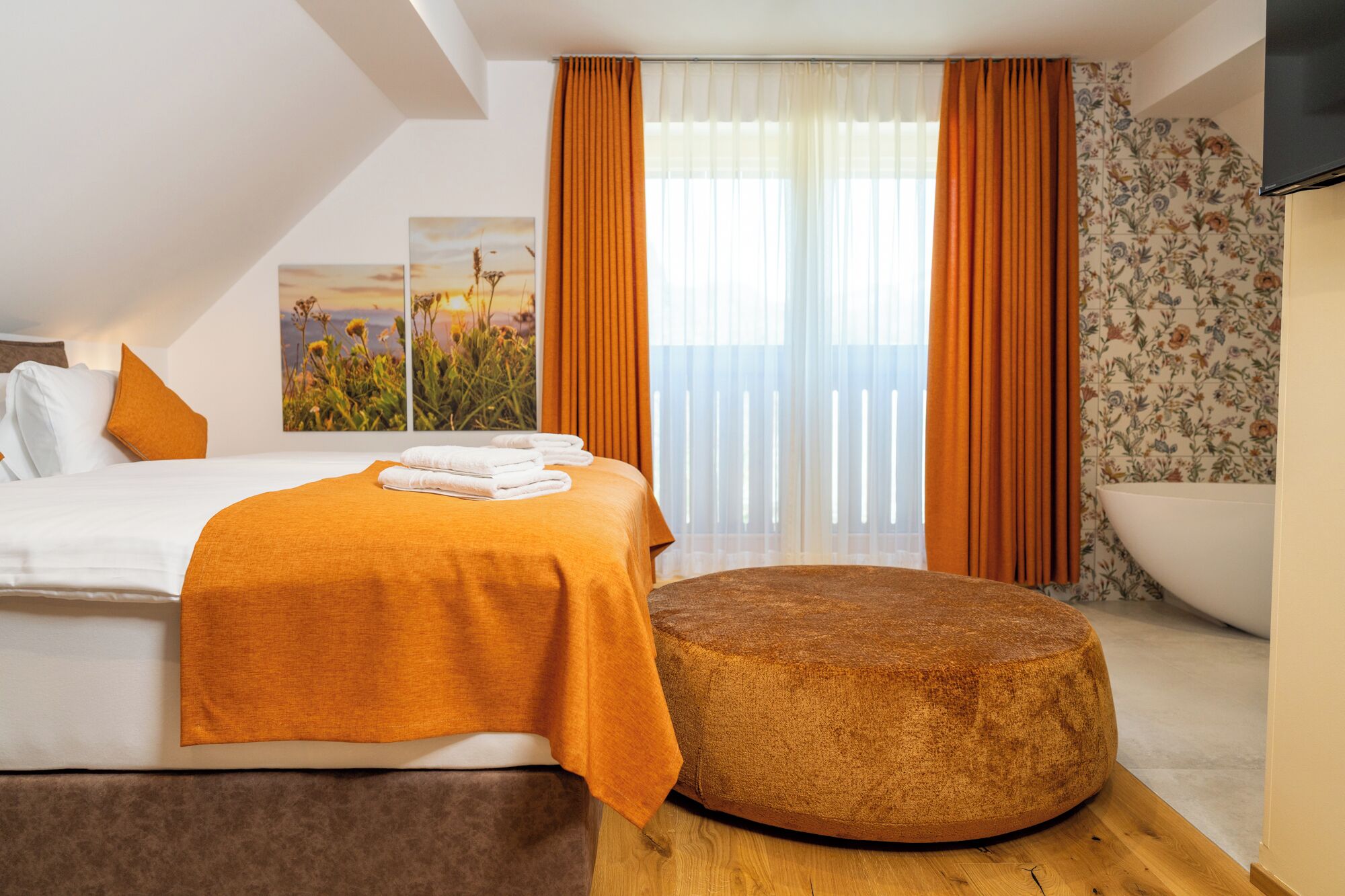 A modern hotel room with a bed, a round ottoman, a large window with curtains, and a view of the sea.
