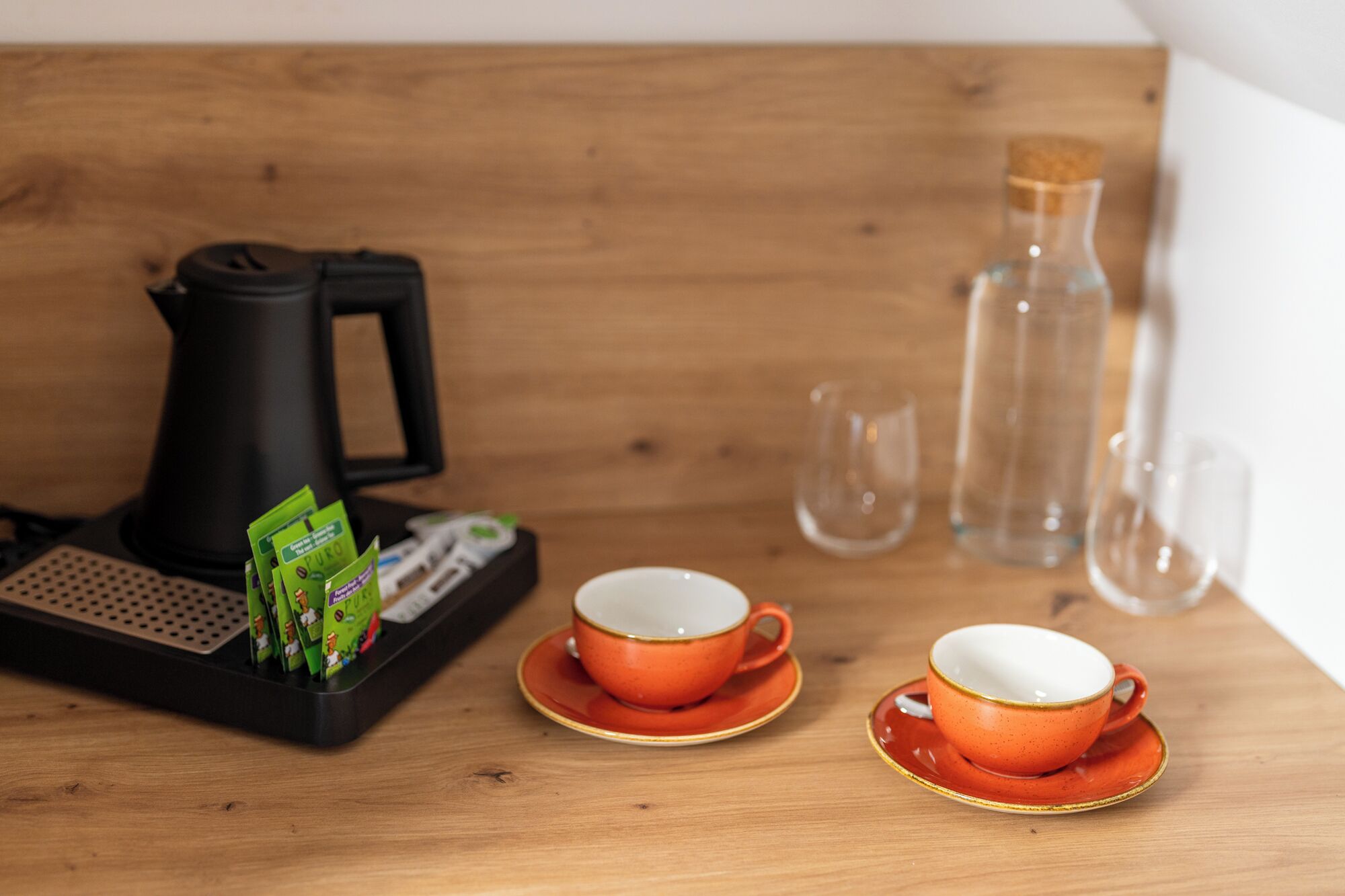 A wooden table with two orange cups on saucers, a black coffee maker, and a glass bottle.