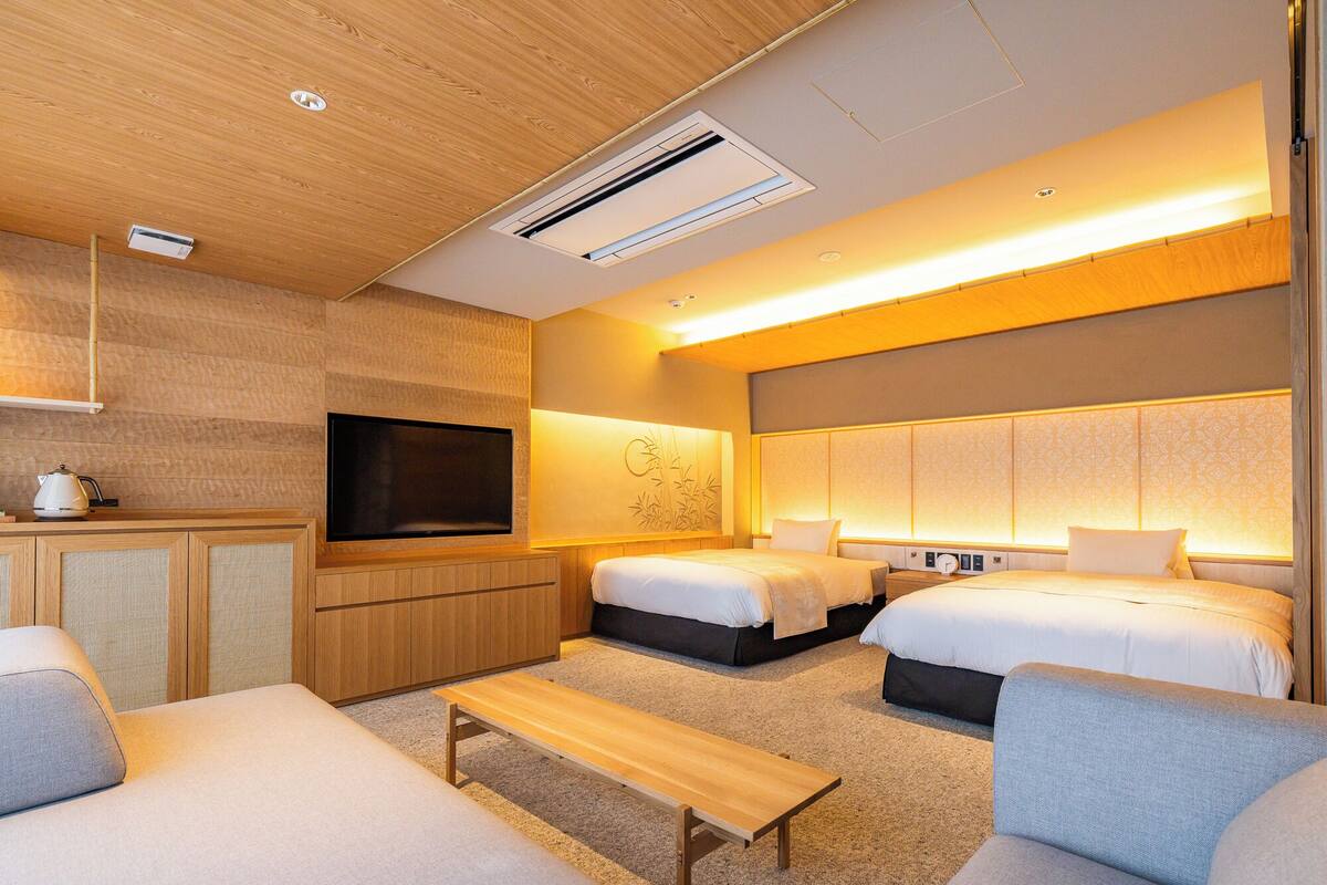 Une chambre d’hôtel moderne avec un plafond en bois, un téléviseur à écran plat, deux lits avec des têtes de lit, un canapé, une petite table et une kitchenette équipée d’une bouilloire.