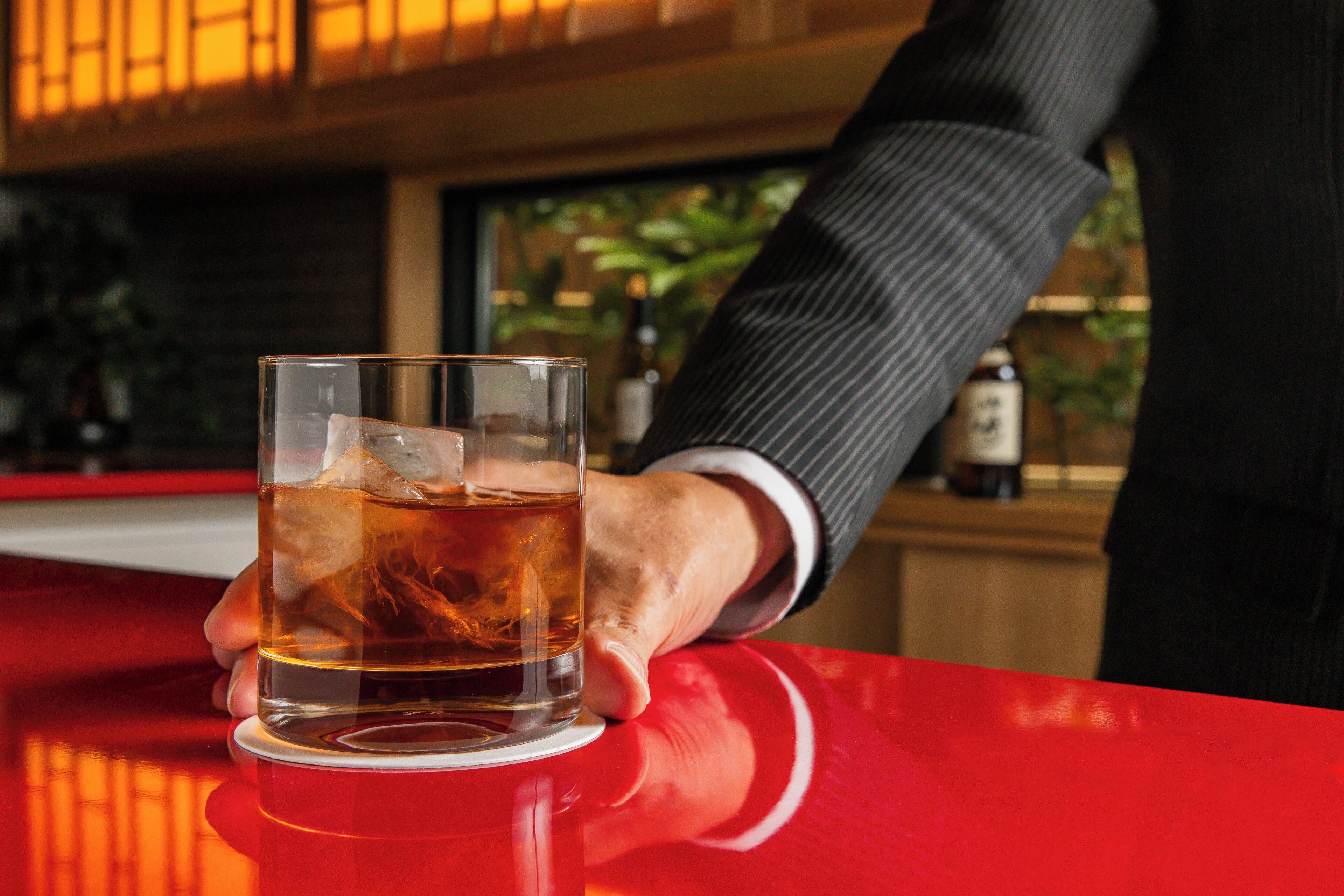 A hand holding a glass of whiskey on a red bar counter.