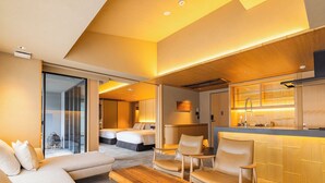 Une chambre d’hôtel moderne comprenant un canapé, deux fauteuils, une table basse en bois, un coin cuisine avec un évier et une cuisinière, et une porte coulissante donnant sur un balcon.