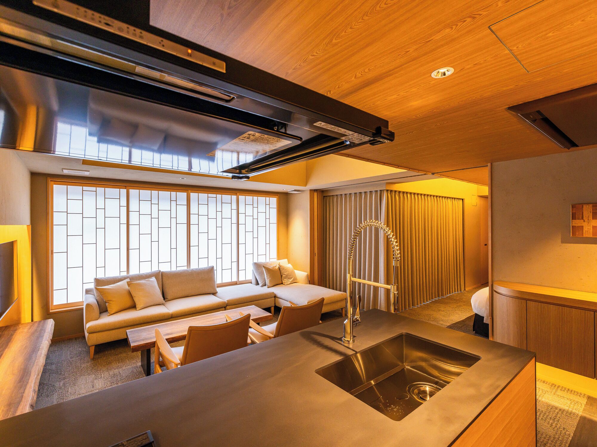 A modern hotel room with a sofa, a dining table, a sink, and a large window with blinds.
