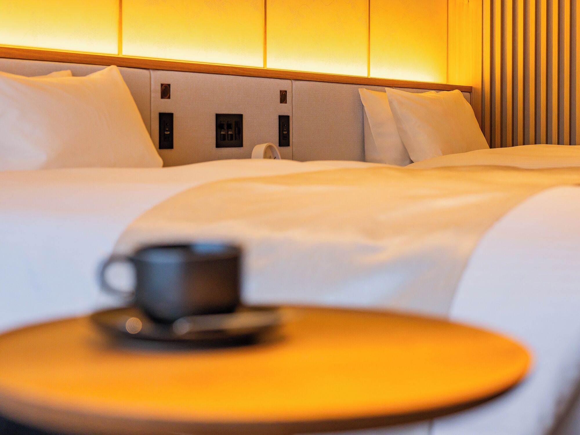 A hotel room with a neatly made bed, a bedside table with a cup, and a wall with vertical blinds.