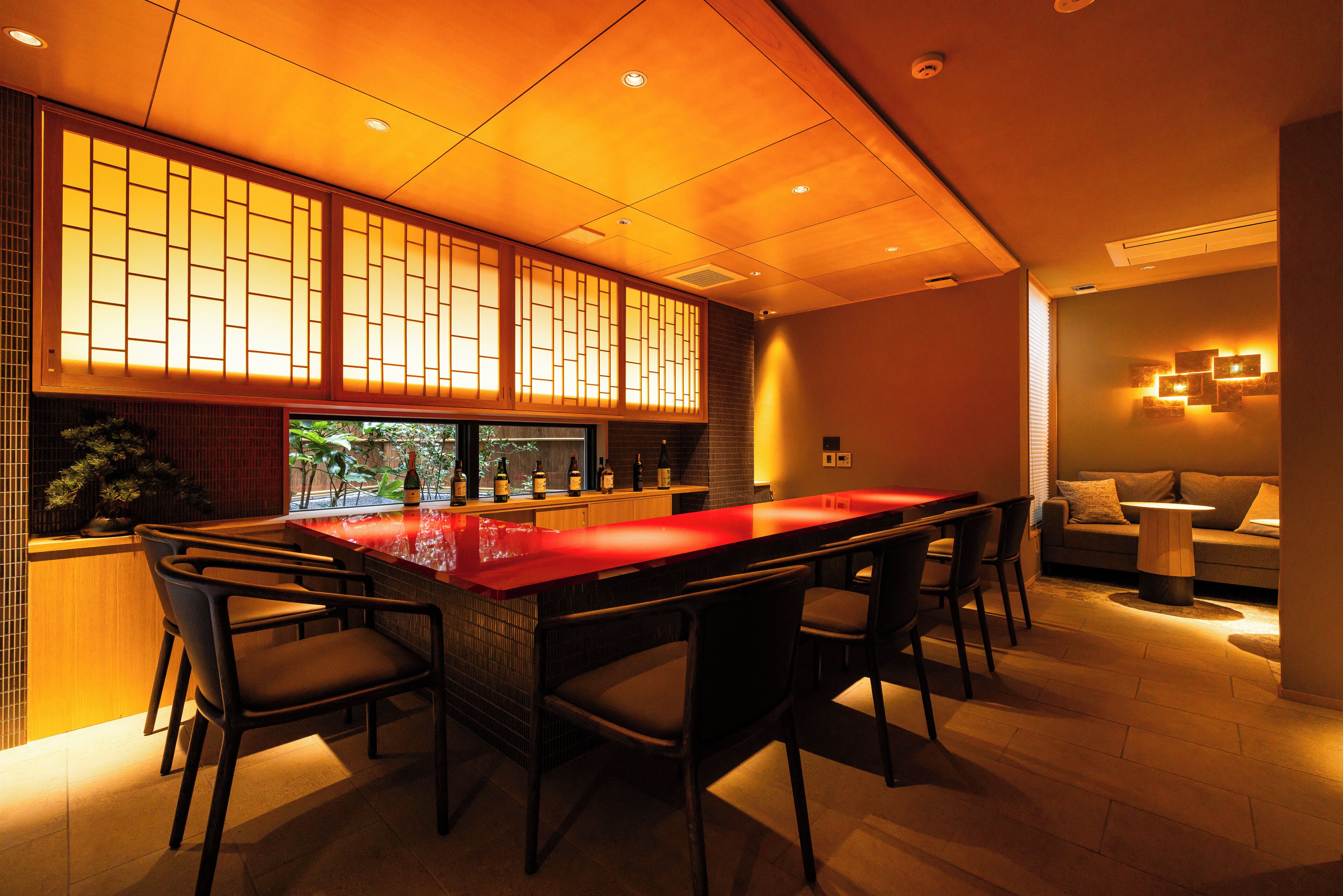 A modern bar area with a long counter, several chairs, and a view of the outside through a large window.