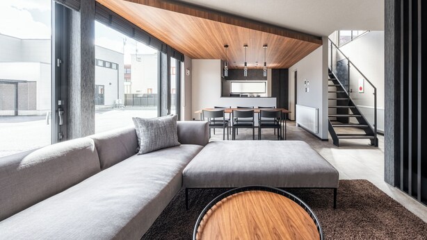 A modern living room with a large sectional sofa, a round coffee table, and a dining area with a table and chairs. A staircase is visible in the background. - JANUS villa Furano (Furano)