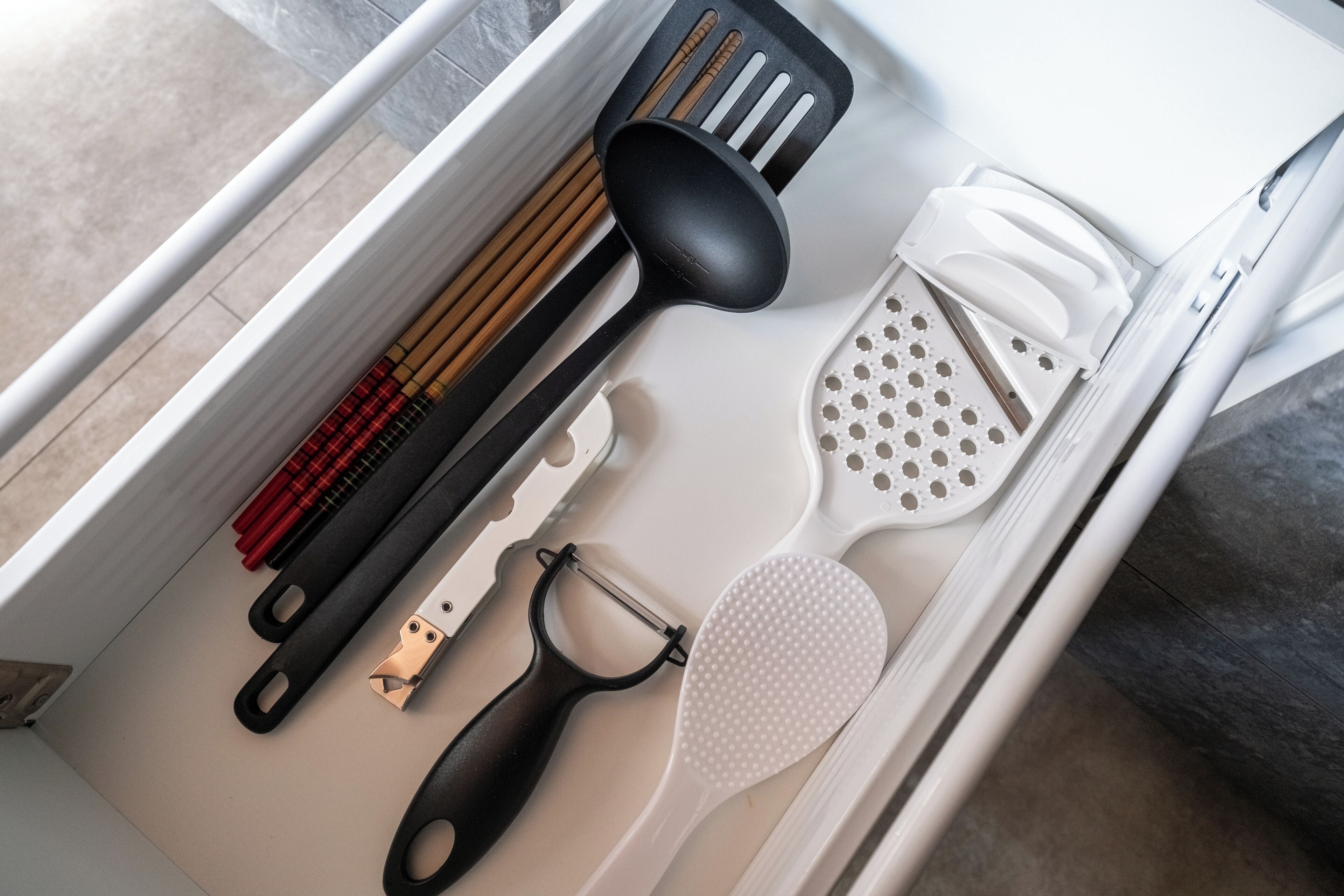 A drawer containing kitchen utensils including a black spatula, a white spatula, a black grater, a white grater, and a pair of wooden chopsticks.