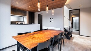 A modern dining area with a wooden table and chairs, a kitchen in the background, and a staircase leading to another level. - JANUS villa Furano (Furano)