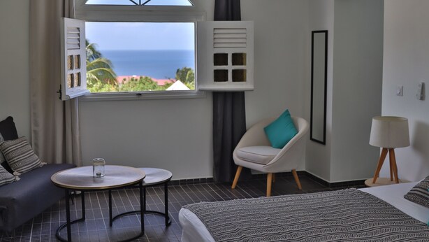A hotel room with a bed, a round table, a chair, a window with a view of the ocean, and a wall-mounted air conditioning unit. - L'Hacienda (Saint-Pierre)