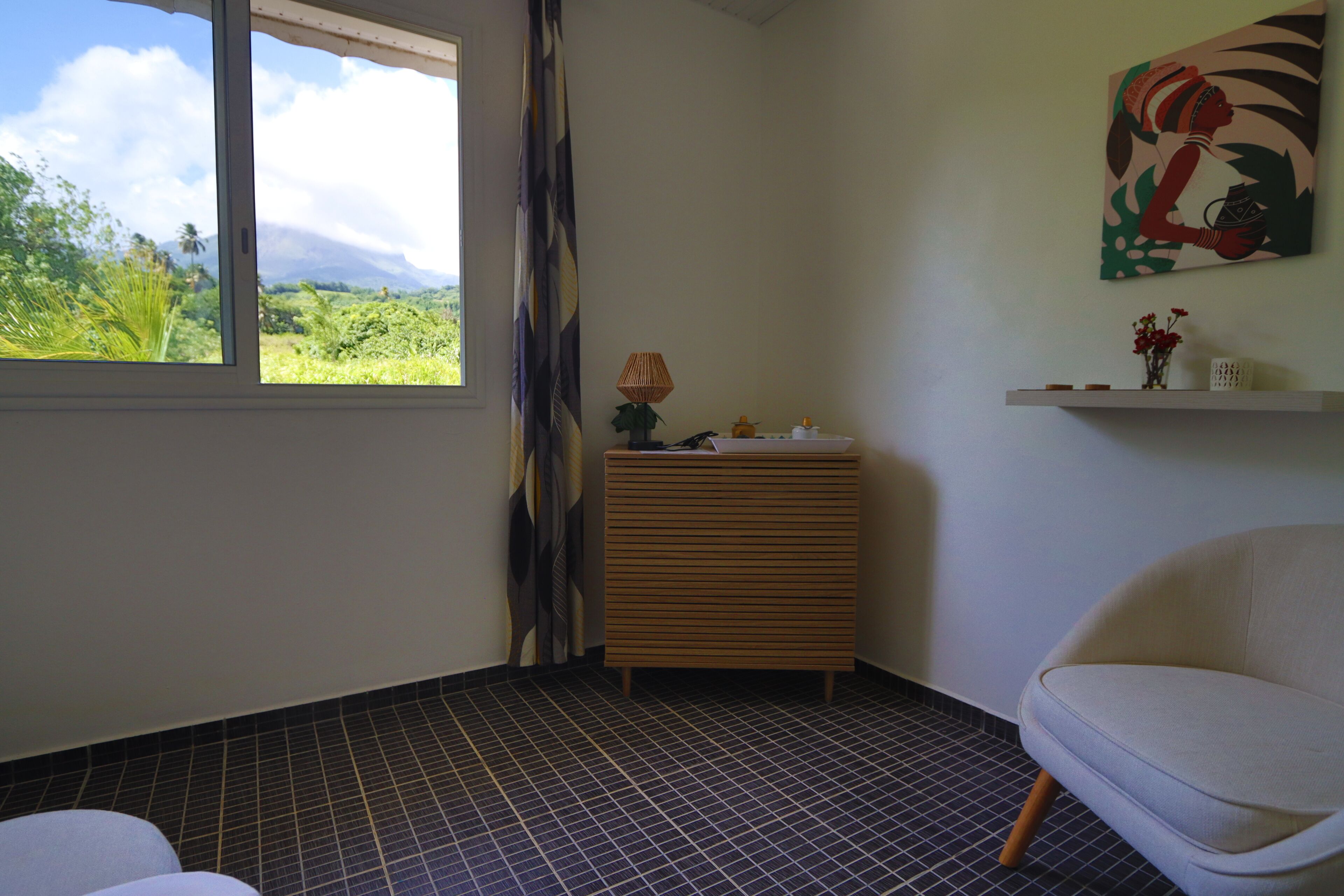 A room with a window, a chair, a small table with a lamp, a shelf with decorative items, and a painting on the wall.