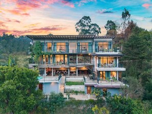 A multi-story house with a balcony on each floor, surrounded by lush greenery.