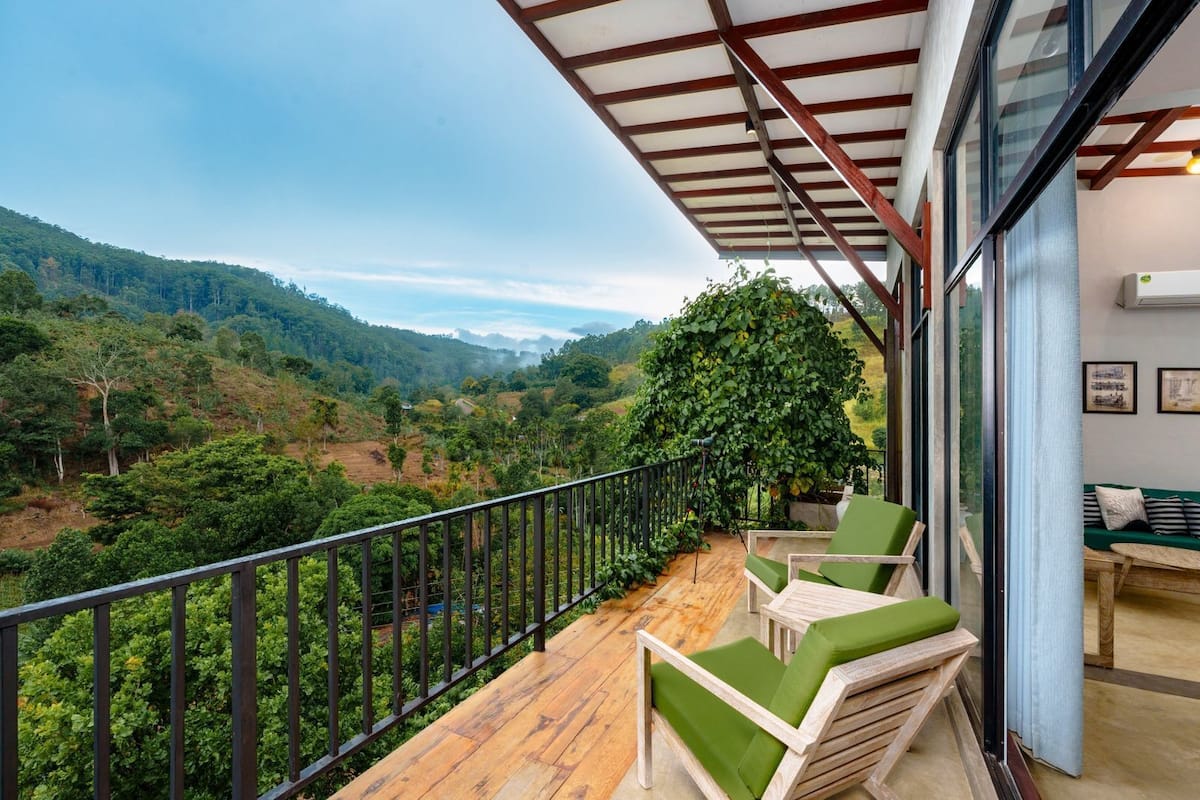 Un balcon doté d’un plancher en bois, de fauteuils confortables aux coussins verts et offrant une vue sur une vallée verdoyante.