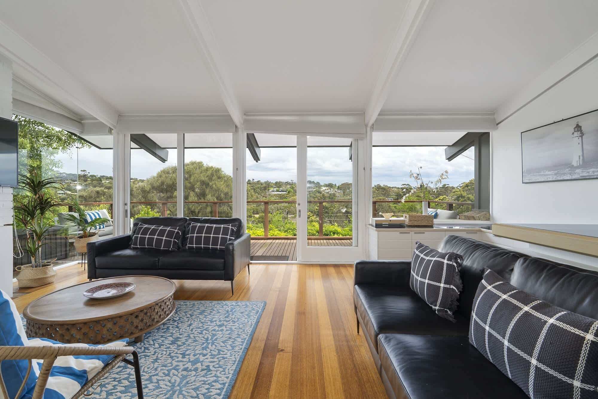 A spacious living room with a black leather sofa, a black armchair, and a round coffee table. There is a large window with a view of greenery and a lighthouse.